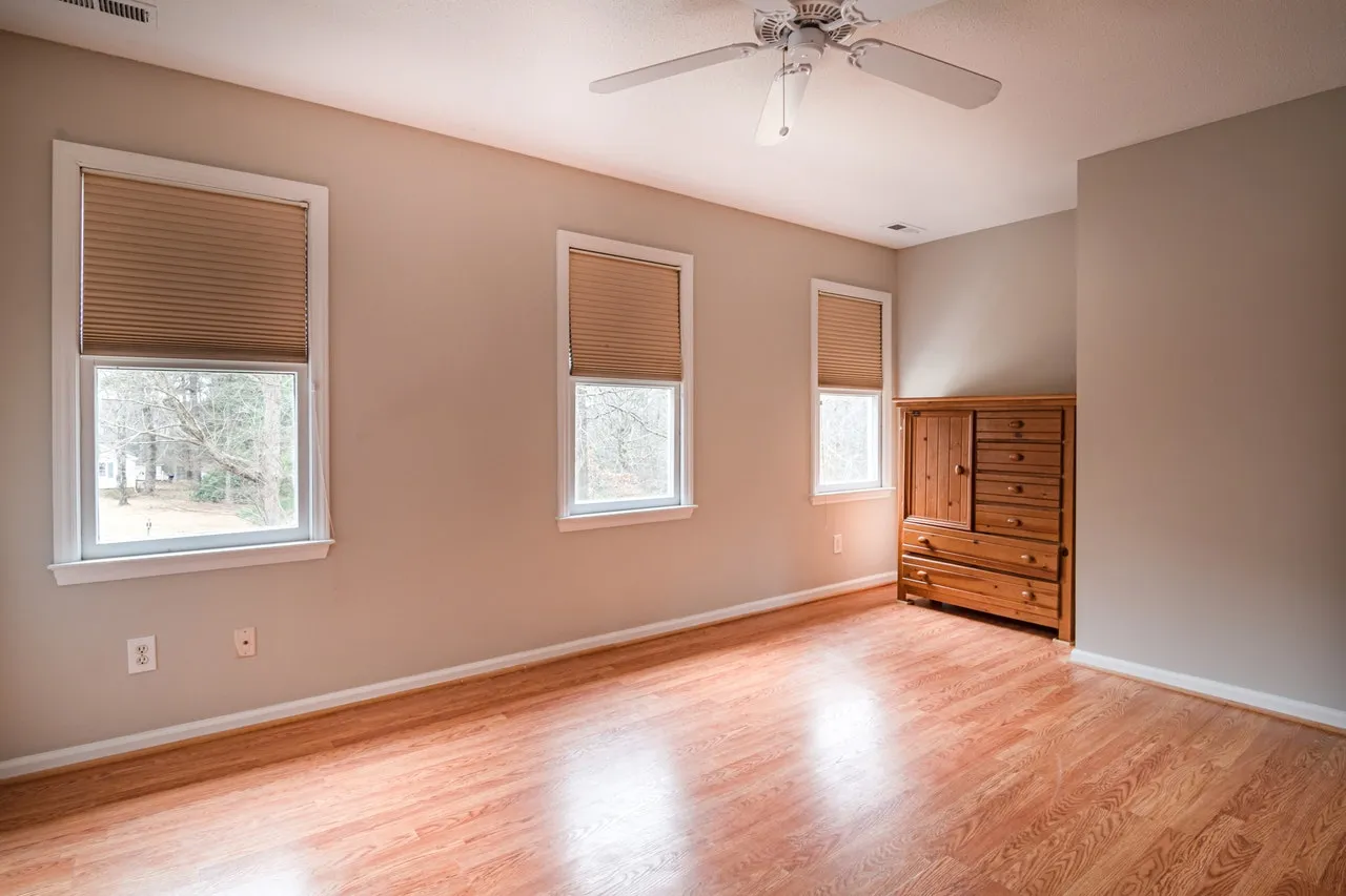 An empty room with a laminate floor, three windows with blinds and a wooden dresser in the corner