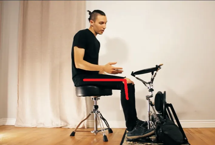 Drummer sitting on a throne in front of an e-drum pedal, a red line shows the thigh sloping down toward the knee